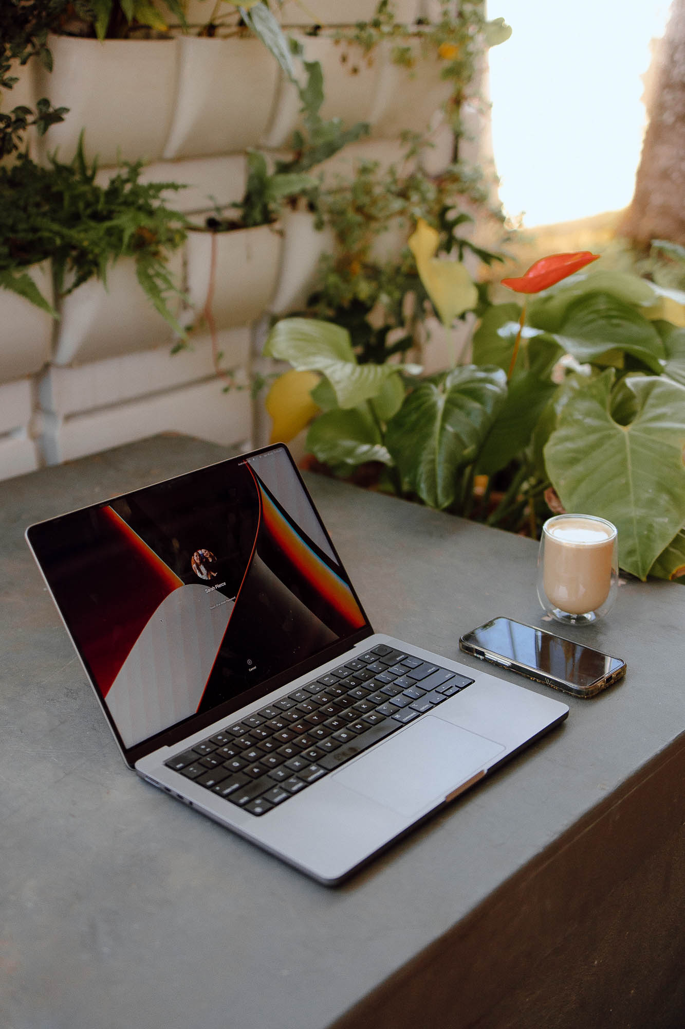 Apple Laptop screen, opened with an abstract black and orange background. A latte and iPhone lie on the concrete table next to the laptop, surrounded by leafy greenery. An ideal outdoor workspace